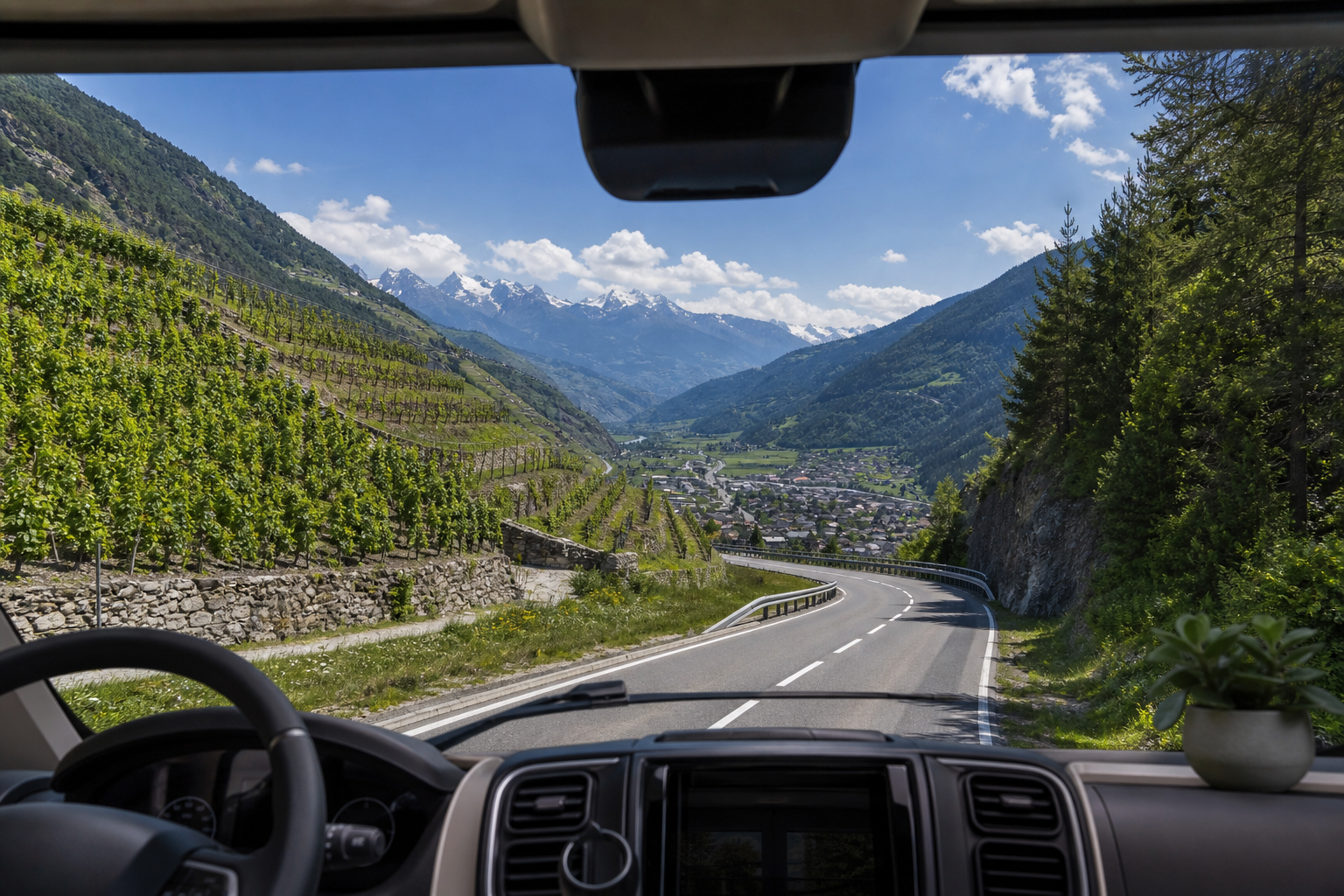Vue depuis le pare-brise d'un camping-car sur une route de montagne sinueuse en Valais, vignobles en terrasses à gauche, versant alpin boisé à droite, village valaisan au loin