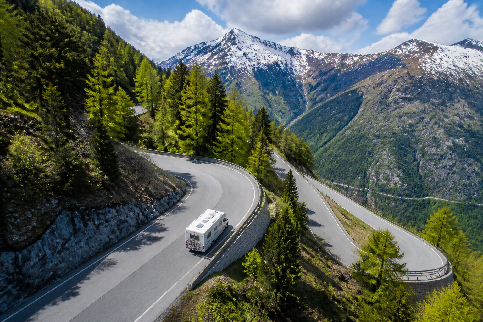 Vue aérienne d'un camping-car blanc sur une route de montagne suisse en lacets, entouré de forêts et de sommets enneigés, sans texte visible